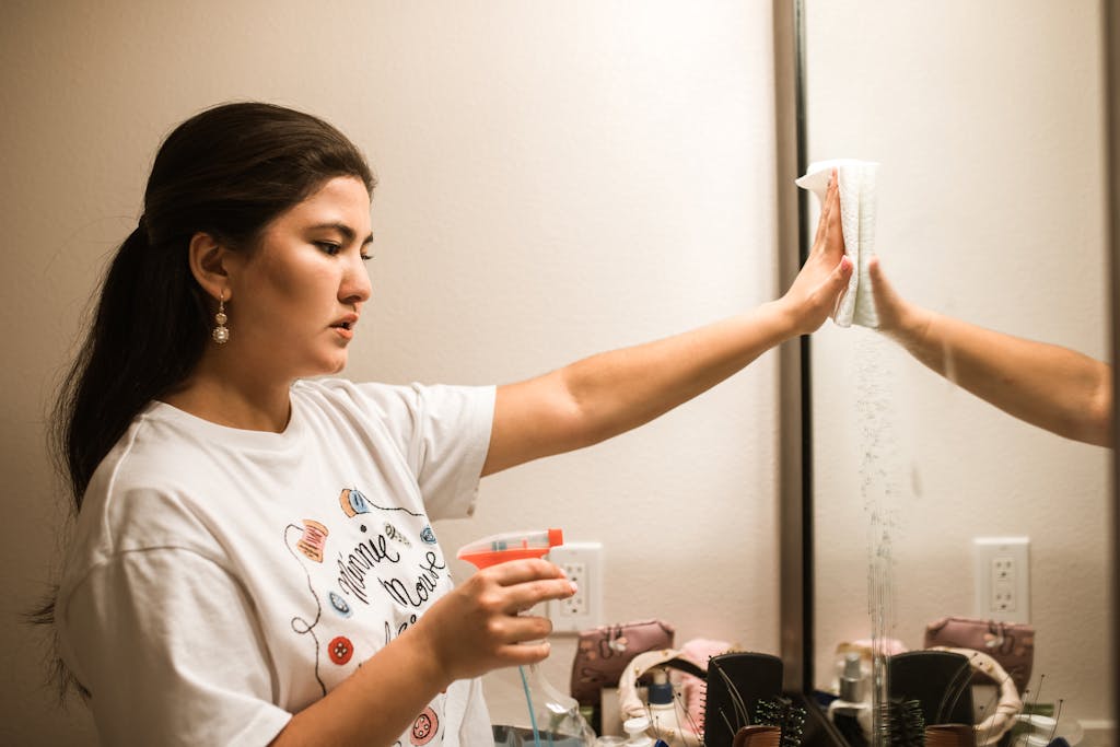 Asian woman using spray bottle to clean mirror. Focus on cleaning chore indoors.