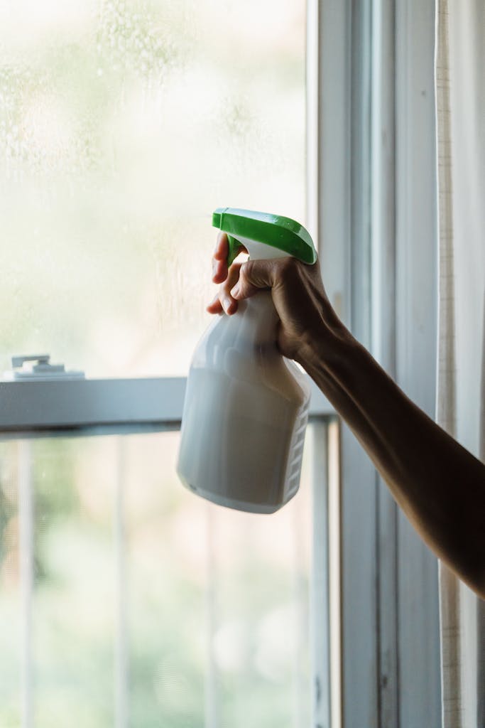 Close-up of hand using a spray bottle for window cleaning indoors, with natural light shining through.