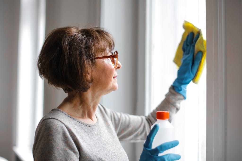 Elderly woman cleansing window indoors using gloves and cleaner.