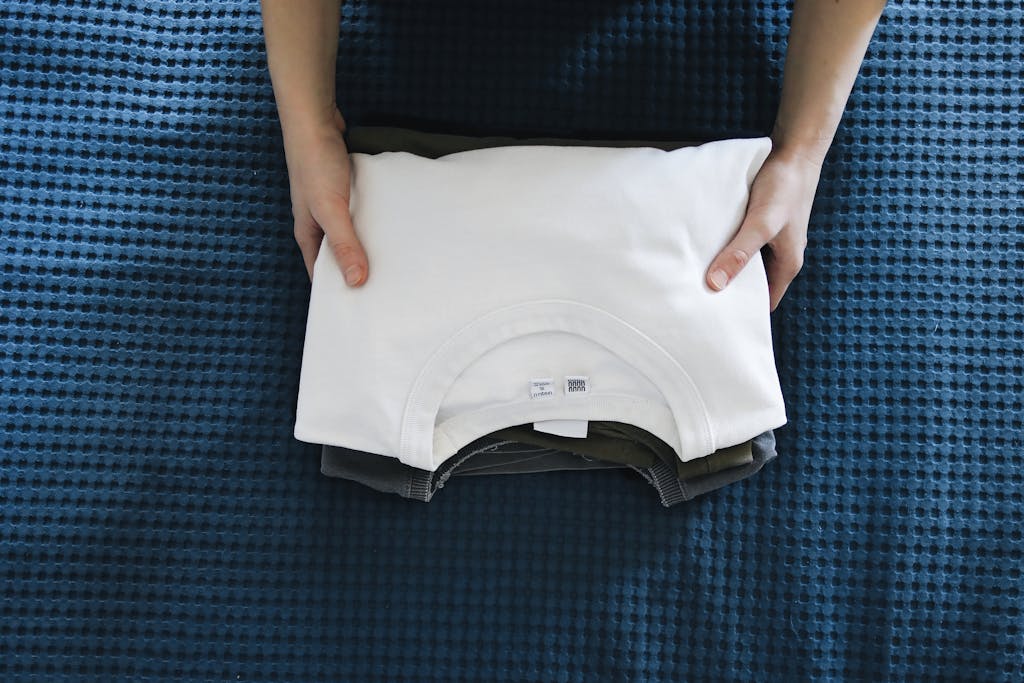 Hands folding a white shirt on a blue textured bedspread, focusing on organizing clothes.