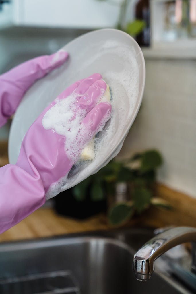 Hands in pink gloves washing a plate with a sponge in a kitchen sink, featuring bubbles and soap.