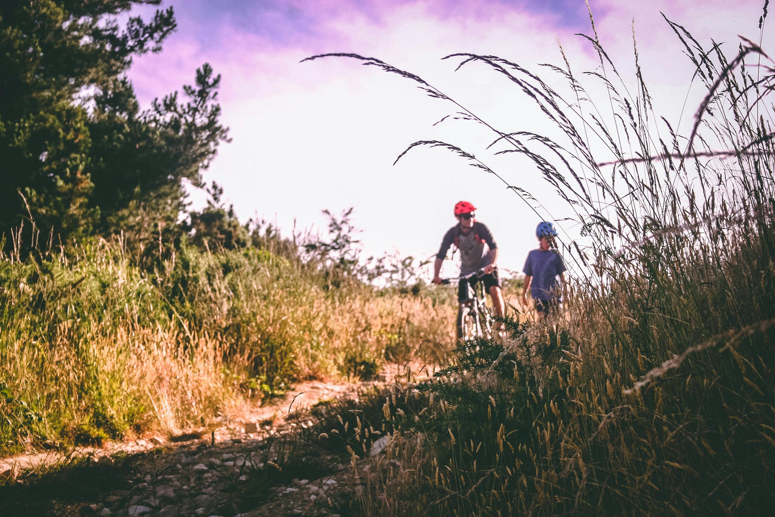 A father and child enjoy mountain biking on an unpaved path surrounded by tall grass and trees.