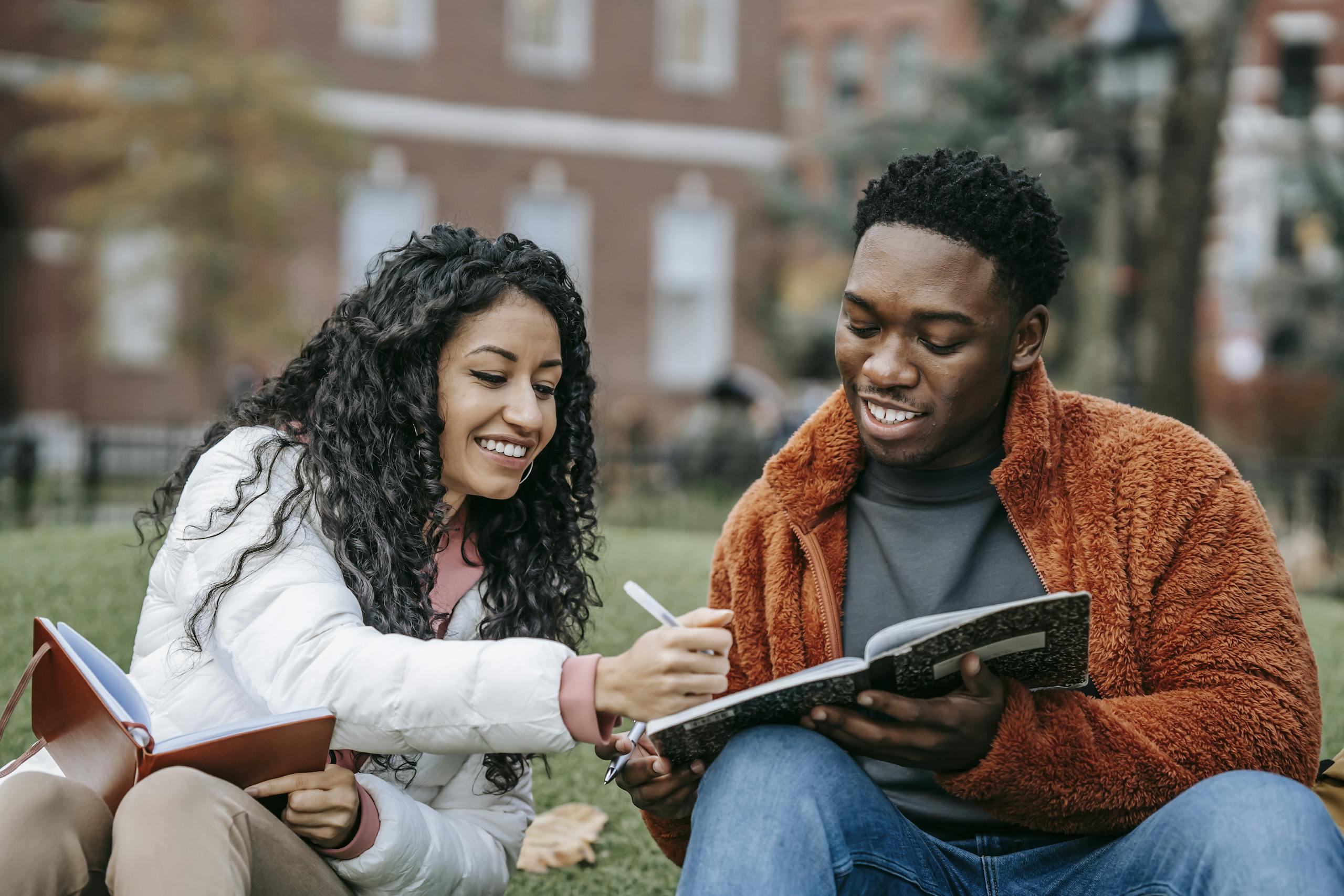 Happy college students studying together on a campus lawn, sharing ideas and writing notes.